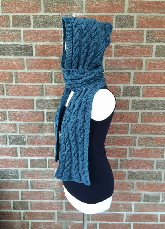 Schaufensterpuppe trägt dunkelblauen gestrickten Kapuzen‑Schal im Zopfmuster vor einer Ziegelwand