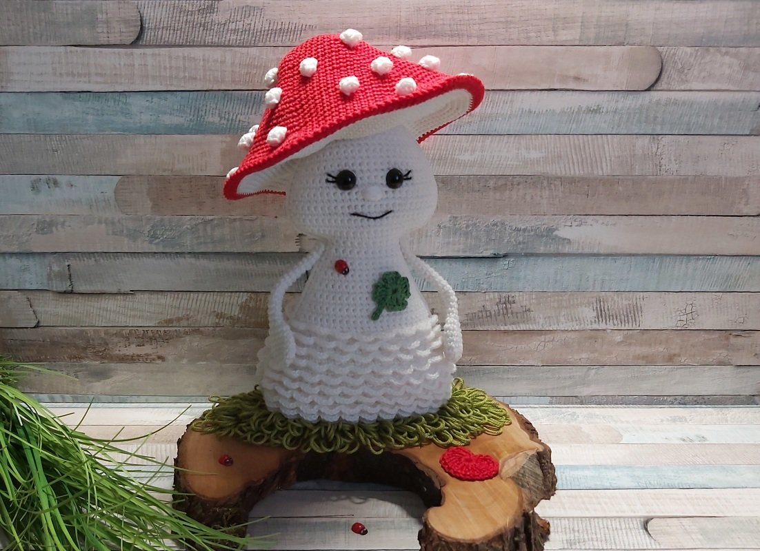 Gehäkelter Fliegenpilz-Amigurumi mit rotem Hut, weißen Punkten, gewelltem Rock und Kleeblatt-Applikation auf Holzscheibe