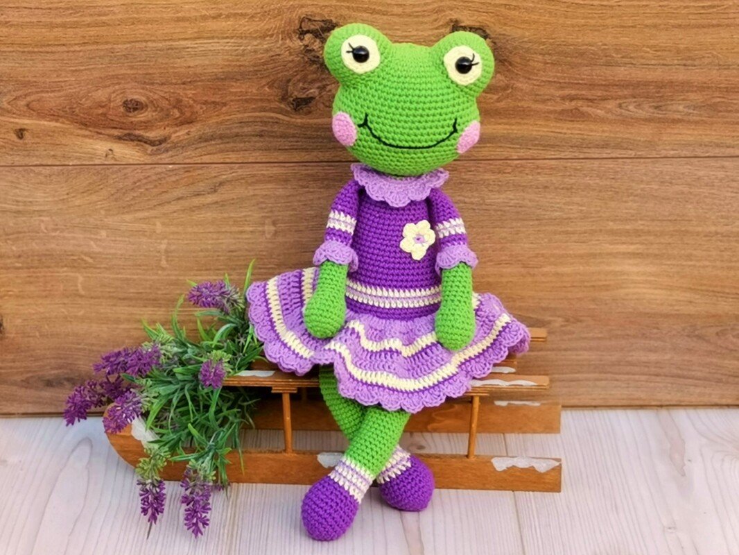 Green crocheted frog in a purple ruffled dress seated on a wooden bench with lavender