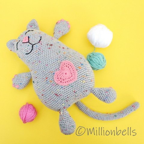 Speckled hand-crocheted amigurumi cat with pink heart applique and yarn balls on yellow background