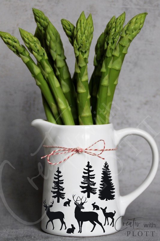 Weißer Keramikkrug mit schwarzen Silhouetten von Hirschen, Rehen, Hasen und Tannenbäumen, gefüllt mit grünem Spargel vor grauem Hintergrund.