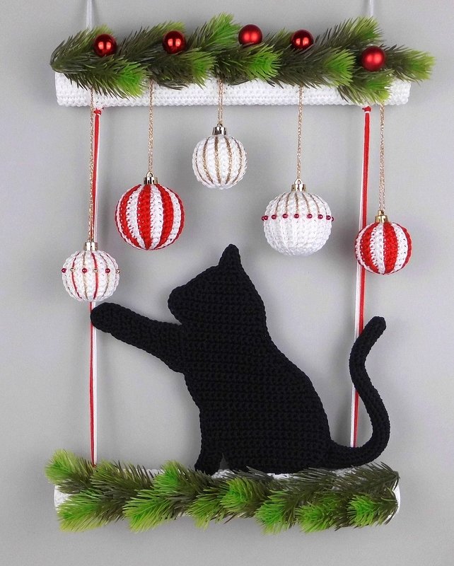 Black crocheted cat reaching for red and white crochet baubles with green evergreen sprigs and small red ornaments.