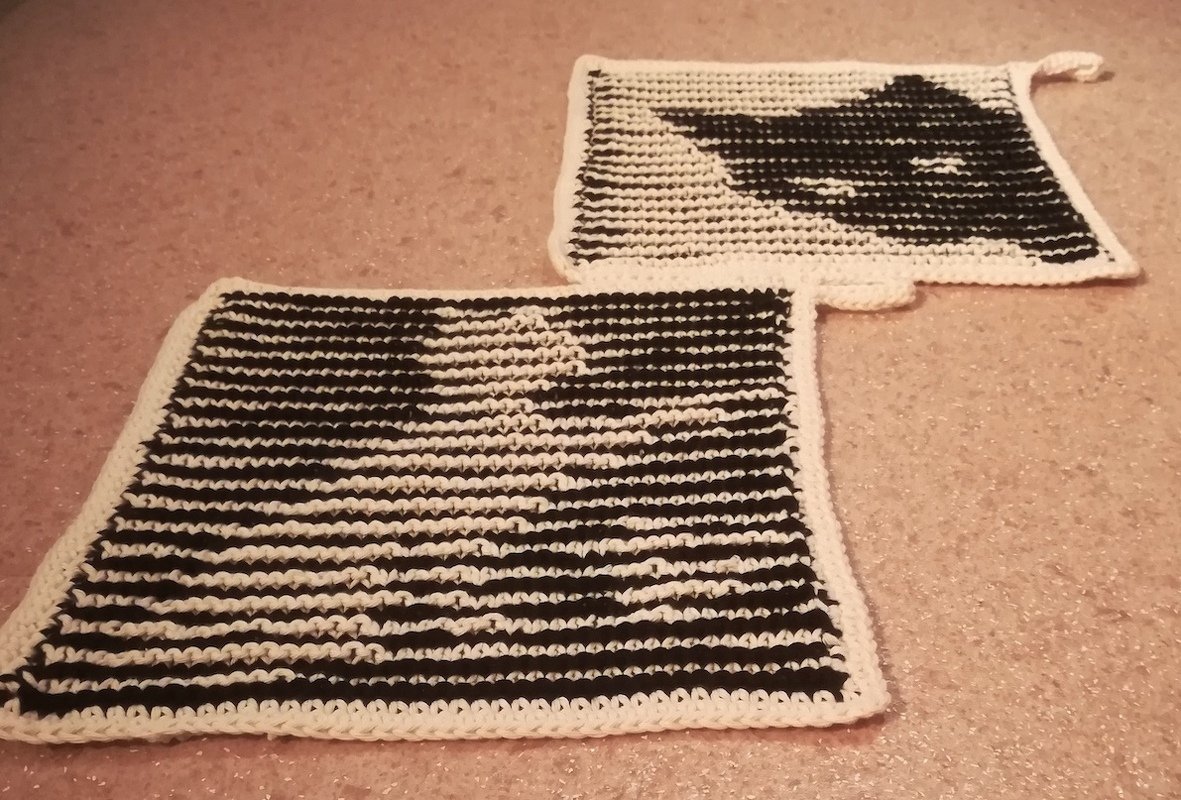 Zwei quadratische schwarz-weiße gehäkelte Topflappen mit Katzen-Silhouette und weißem Rand liegend