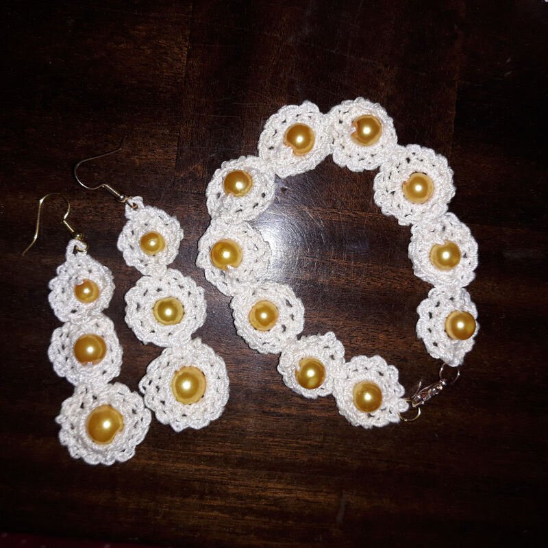 Weiße gehäkelte Blütenohrringe und passendes Armband mit goldfarbenen Perlen auf dunklem Holz.