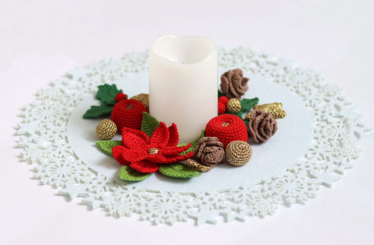 Handgehäkelte Weihnachtstischdeko mit rotem Weihnachtsstern, Tannenzapfen, goldenen Kugeln und einer weißen Kerze auf einem Schneeflocken-Deckchen.