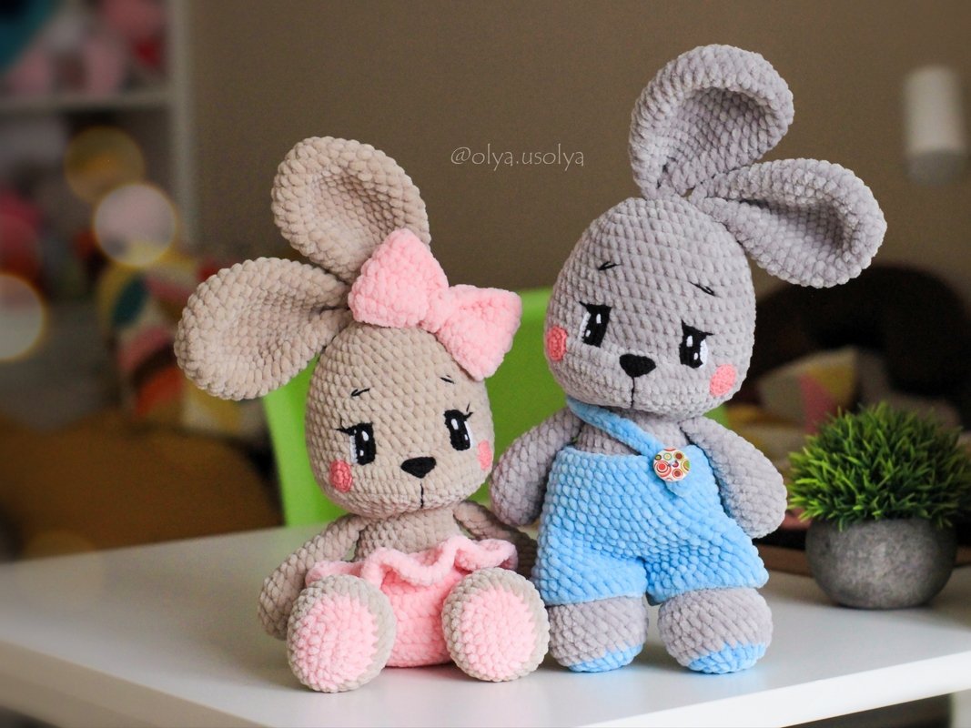 Two crocheted amigurumi bunnies, one beige with a pink bow and skirt and one grey in blue overalls, seated on a white surface.
