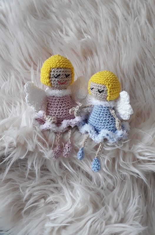 Zwei gehäkelte Amigurumi-Engelchen mit gelben Haaren, weißen Flügeln und Kleidern in Rosa und Hellblau auf flauschigem Untergrund.