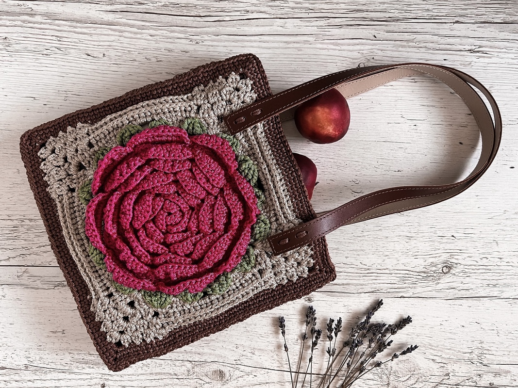 Square crochet shoulder bag with a large pink crocheted rose, brown straps, and two peaches visible nearby on whitewashed wood.