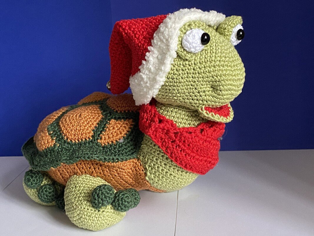 Gehäkelte Amigurumi-Schildkröte mit rot-weißer Weihnachtsmütze und rotem Schal vor blauem Hintergrund