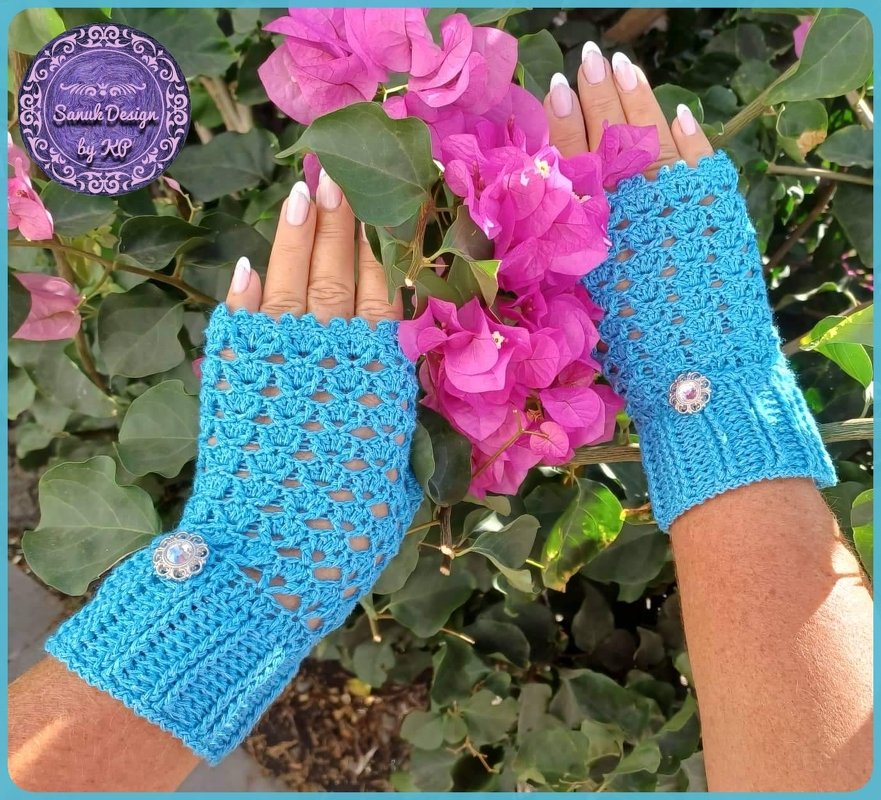 Gehäkelte türkisfarbene fingerlose Handschuhe mit Spitzenmuster und Zierknopf an Damenhänden vor pinken Blüten