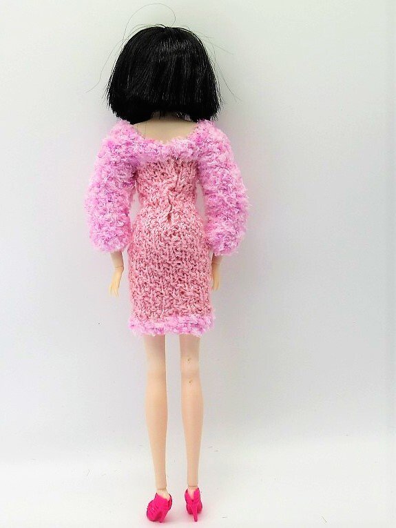 Rückansicht einer Puppe mit kurzem schwarzem Haar, trägt ein rosa gestricktes Kleid mit flauschigen Ärmeln und pinken Schuhen.