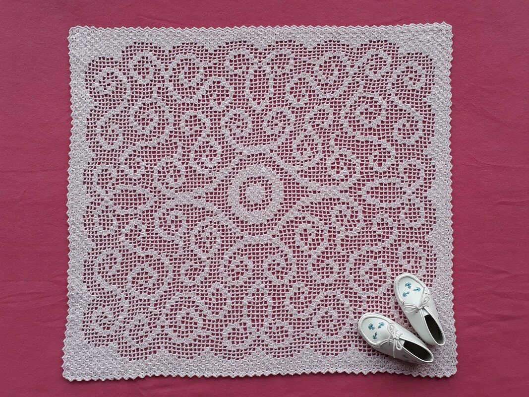 Quadratische weiße Filet-Häkeldecke mit zentralem Rundmotiv auf rosa Hintergrund, daneben weiße Babyschuhe.