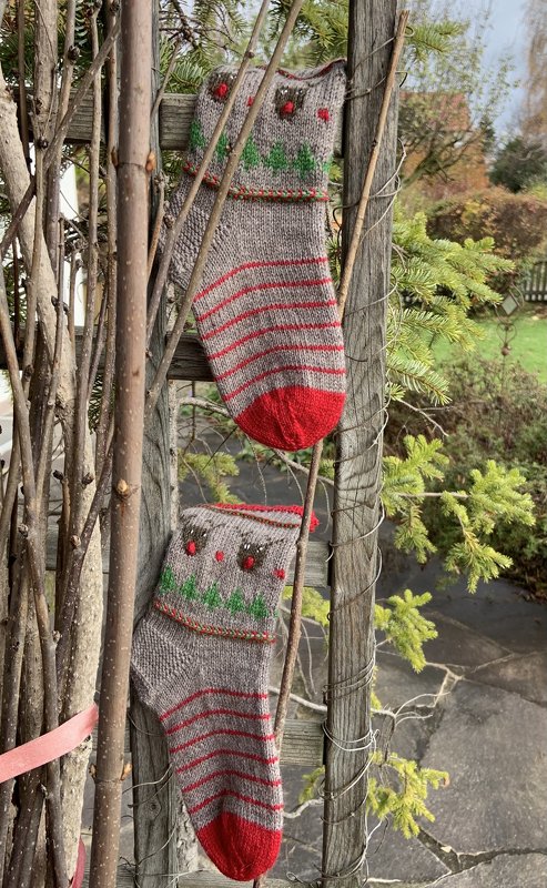 Zwei gestrickte graue Weihnachtssocken mit roten Streifen, roten Spitzen und Rentier‑/Tannenbaum‑Motiv an einem Holzspalier.
