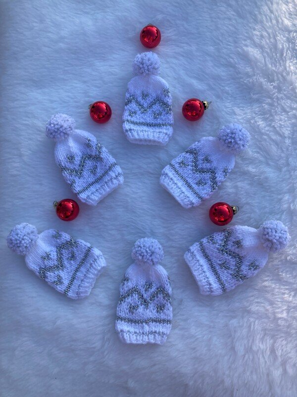 Sieben weiße gestrickte Mini-Mützen mit Bommel und silbernem Zickzackmuster auf weißem Plüsch, dekoriert mit roten Kugeln