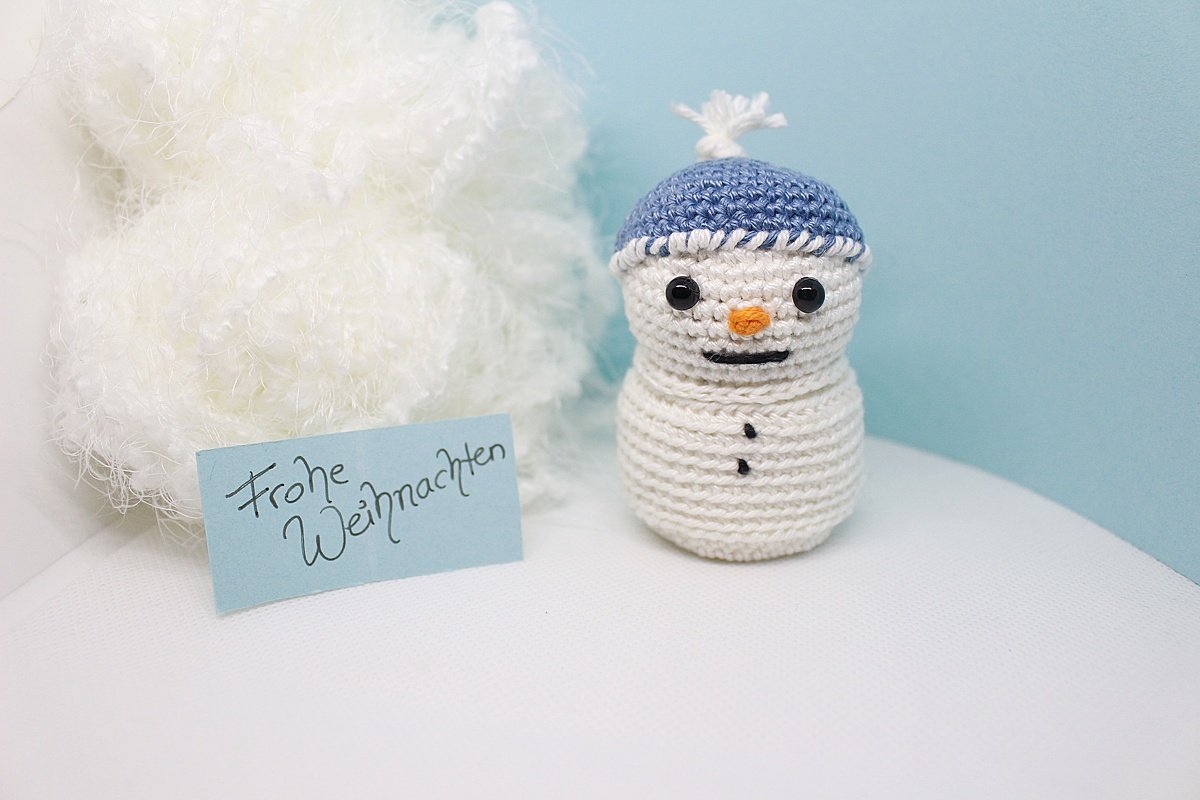 Gehäkelter Schneemann mit blauer Mütze, orangefarbener Nase und Karte mit 'Frohe Weihnachten' vor weißem Flausch