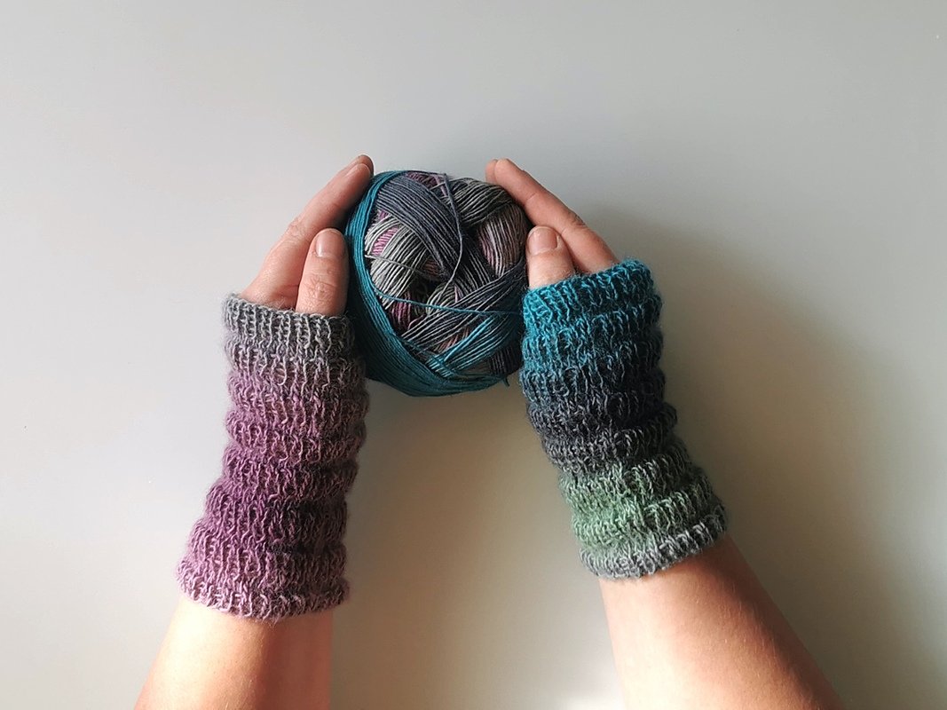 Hände mit fingerlosen Armstulpen aus Verlaufsgarn (Lila bis Grün), Maschenstruktur gut sichtbar, halten ein mehrfarbiges Garnknäuel