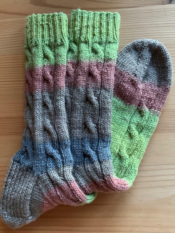 Gestricktes Sockenpaar mit Zopfmuster und pastellfarbenen Streifen auf Holzuntergrund