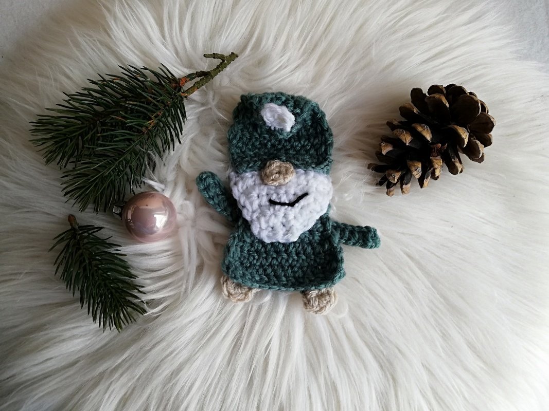 Gehäkelter grüner Wichtel mit weißem Bart, umgeben von Tannenzweigen, Weihnachtskugel und Tannenzapfen auf weißem Fell