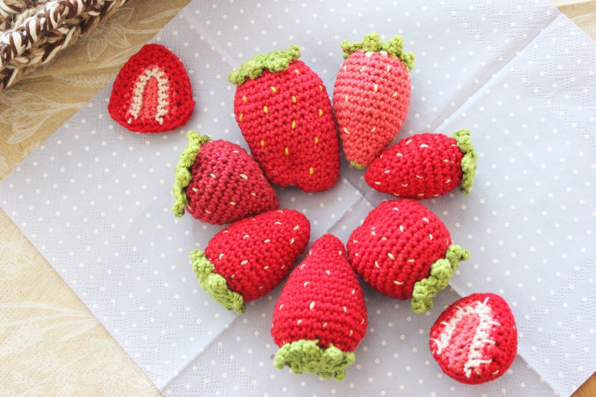 Gehäkelte Erdbeeren in Rot mit grünen Blättern, einige ganze und zwei halbierte, auf hellblauem gepunktetem Stoff.
