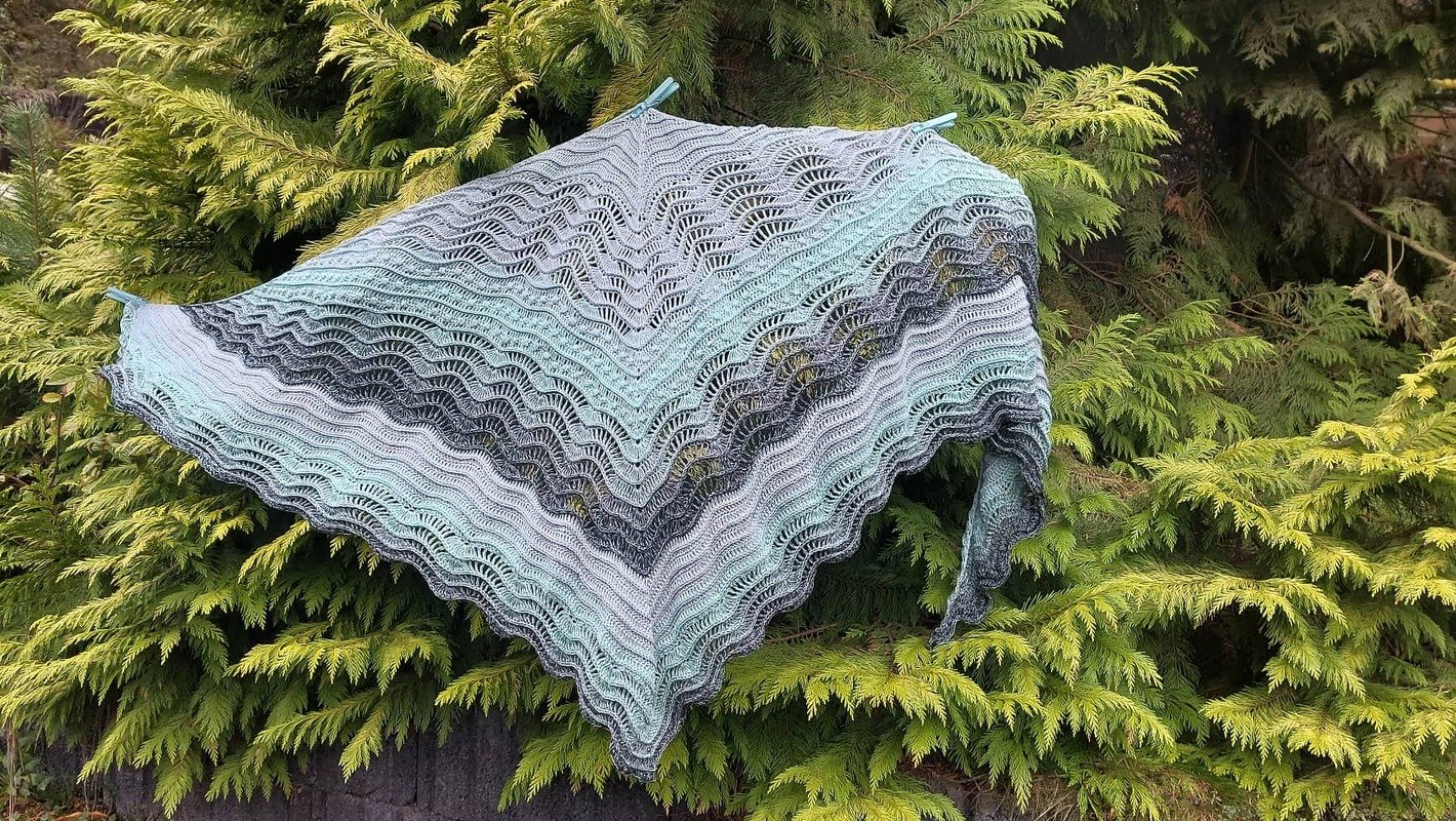 Gehäkeltes dreieckiges Tuch in Mint- und Grautönen mit Wellen- und Spitzendesign, an Wäscheklammern vor grünen Zweigen aufgehängt.
