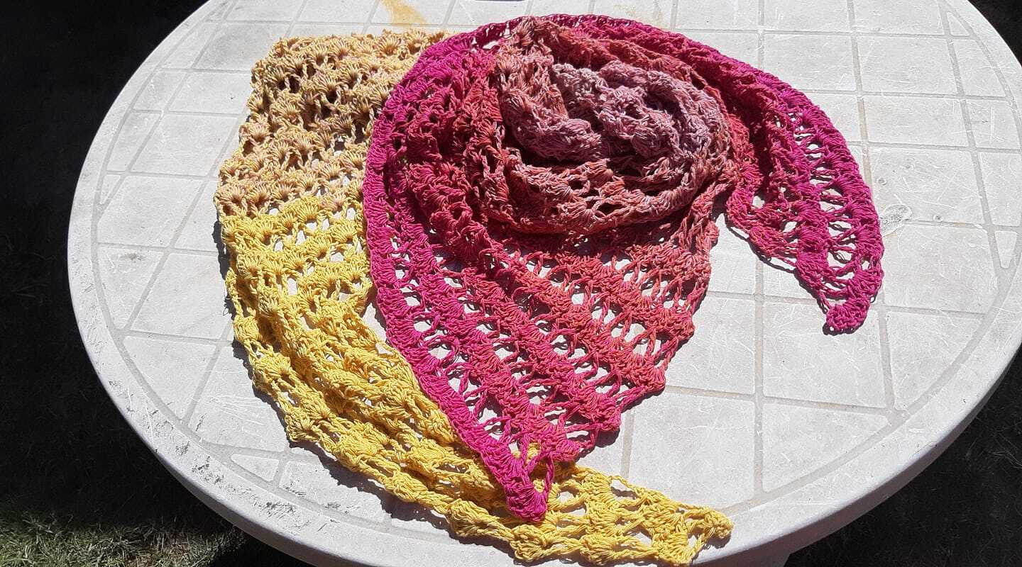 Zwei gehäkelte Dreieckstücher in Gelb und Pink auf rundem Tisch