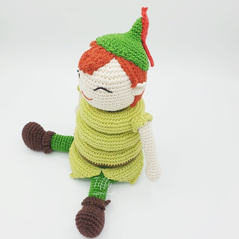 Amigurumi boy doll with orange yarn hair, green tunic, pointed hat with red feather, and brown boots in a sitting pose