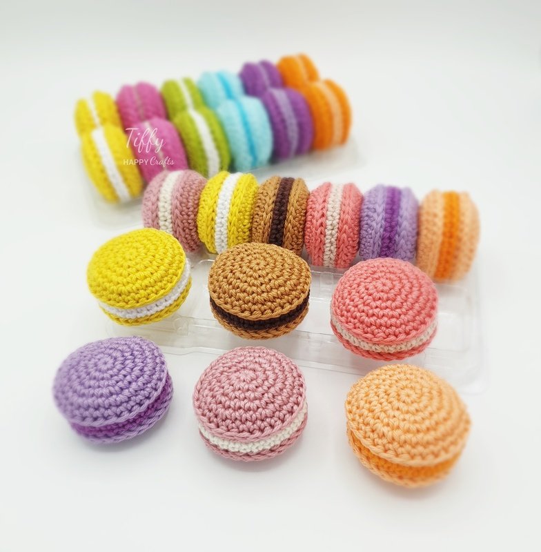 Bright crocheted macarons in a clear organizer and scattered on a white surface, showing color variety and handmade stitch details.
