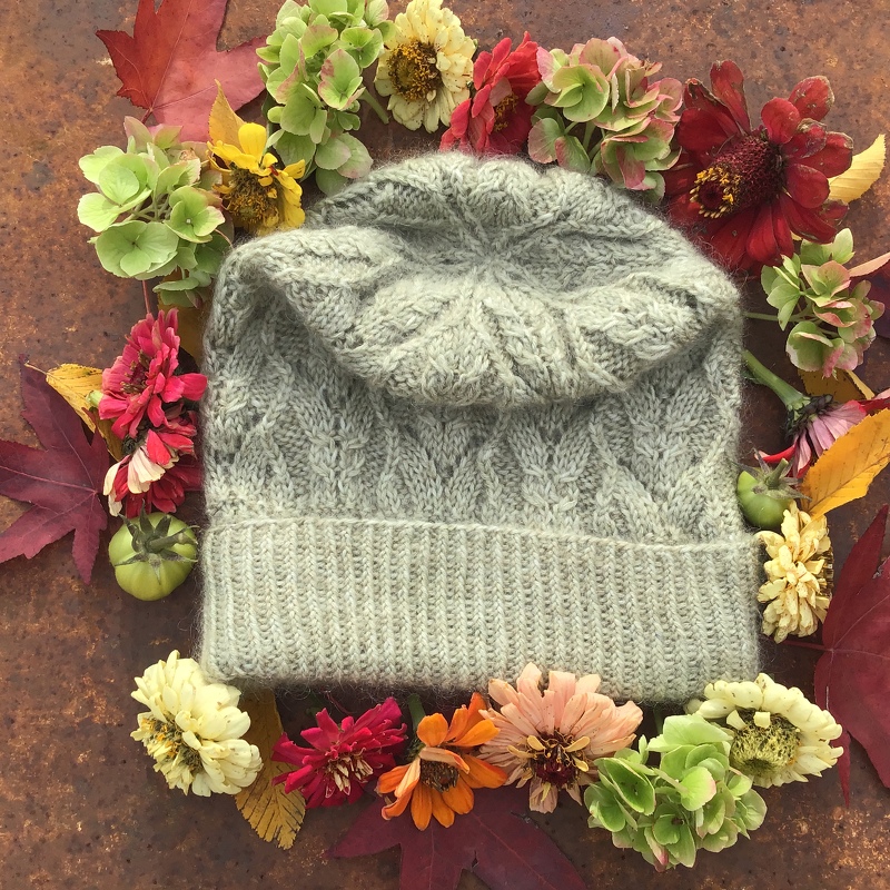 Hellgraue gestrickte Damenmütze mit Blattmuster und umgeschlagenem Bund, auf rostigem Untergrund umgeben von Herbstblumen