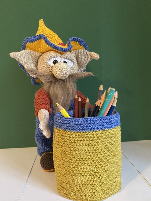 Amigurumi-Zwerg mit gelb-blauem Hut und langem Bart neben gelbem Häkel-Utensilo mit blauem Rand und Buntstiften.