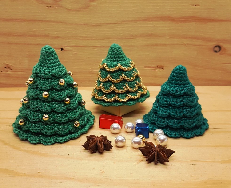Drei schnell gehäkelte Mini-Tannen in Grün, verziert mit goldenen Perlen und Kordel, auf Holzuntergrund mit Perlen, Geschenkschachteln und Sternanis.