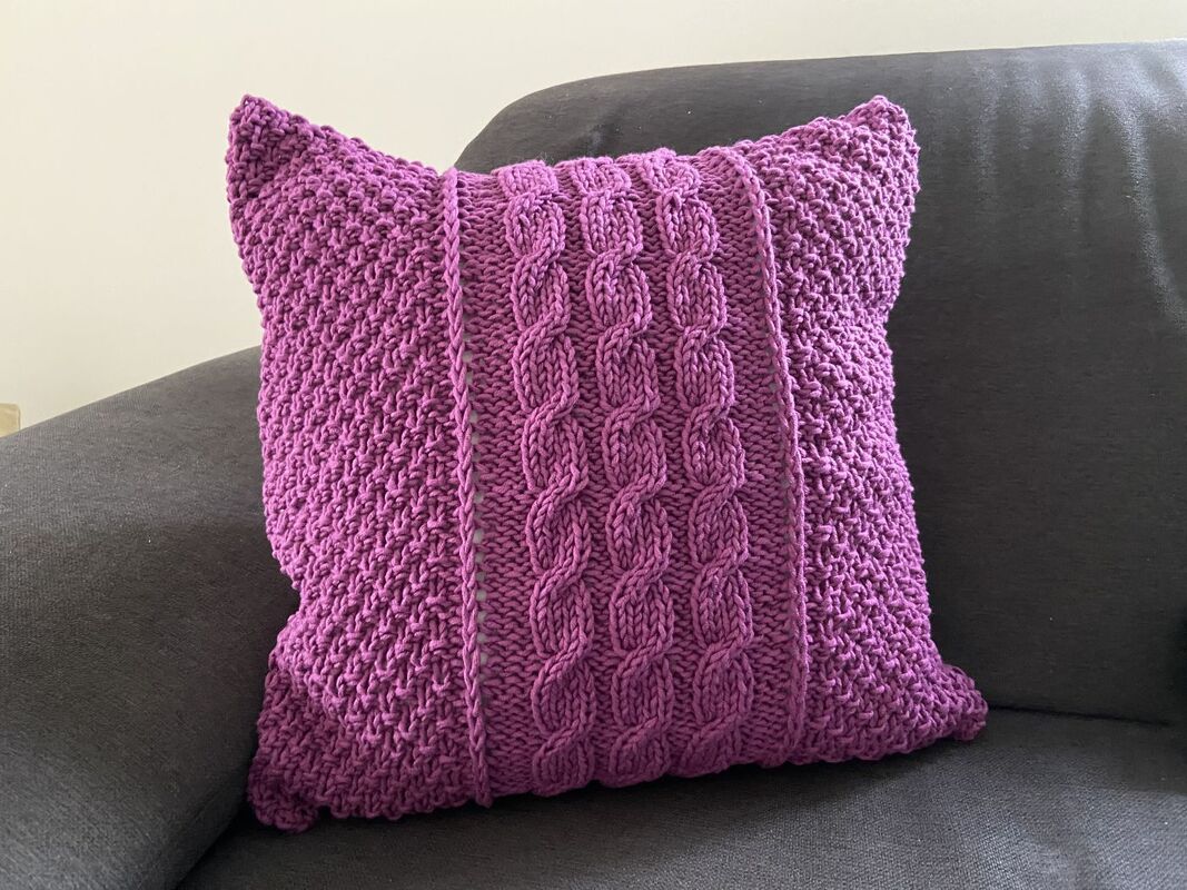 Violettes gestricktes Kissen mit mittigem Zopfmuster auf dunklem Sofa