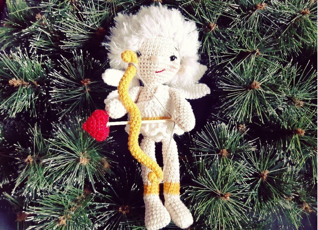 Gehäkelter weißer Amigurumi‑Engel mit gelbem Bogen und rotem Herzpfeil vor Tannenzweigen