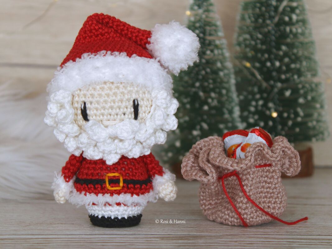 Gehäkelter Weihnachtsmann‑Amigurumi mit beigefarbenem Säckchen vor Mini‑Tannenbäumen