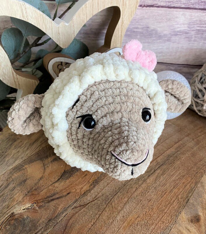 Gehäkelter Amigurumi-Schafkopf mit cremefarbenem Fell, beigem Gesicht und rosa Herz, auf Holztisch liegend.