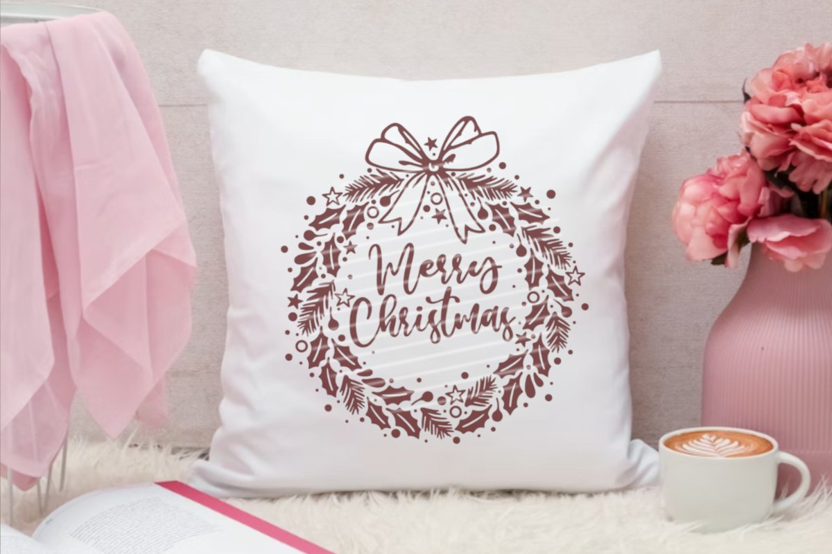 Weißes Kissen mit braunem Plott eines Weihnachtskranzes und dem Schriftzug 'Merry Christmas', daneben rosa Dekoration und eine Tasse Kaffee.