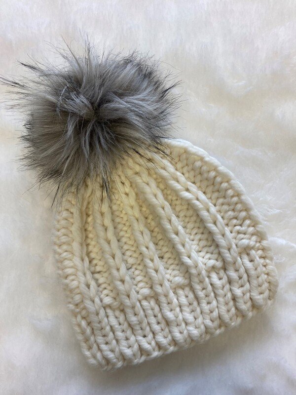 Cremefarbene grob gestrickte Mütze mit großem grauem Bommel auf hellem Hintergrund
