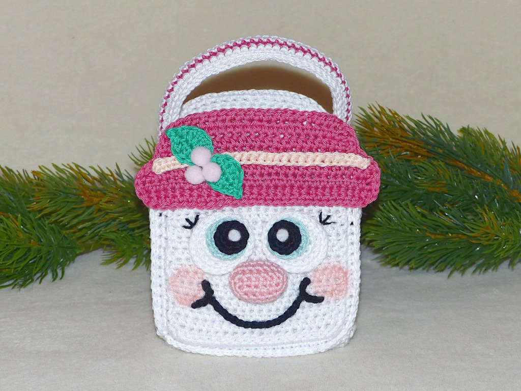 Weißes gehäkeltes Schneemann-Utensilo mit Henkel und pinkem Hut vor Tannenzweigen.