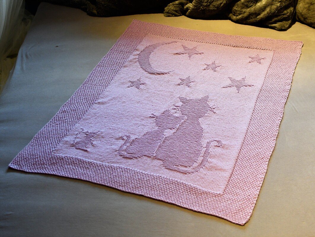 Soft pink knitted baby blanket with two cat silhouettes, a crescent moon, and stars.