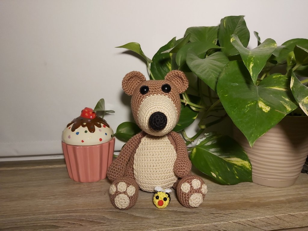 Sitzender gehäkelter Amigurumi-Teddybär in Braun mit hellem Bauch und Pfotenballen, davor eine kleine gehäkelte Biene