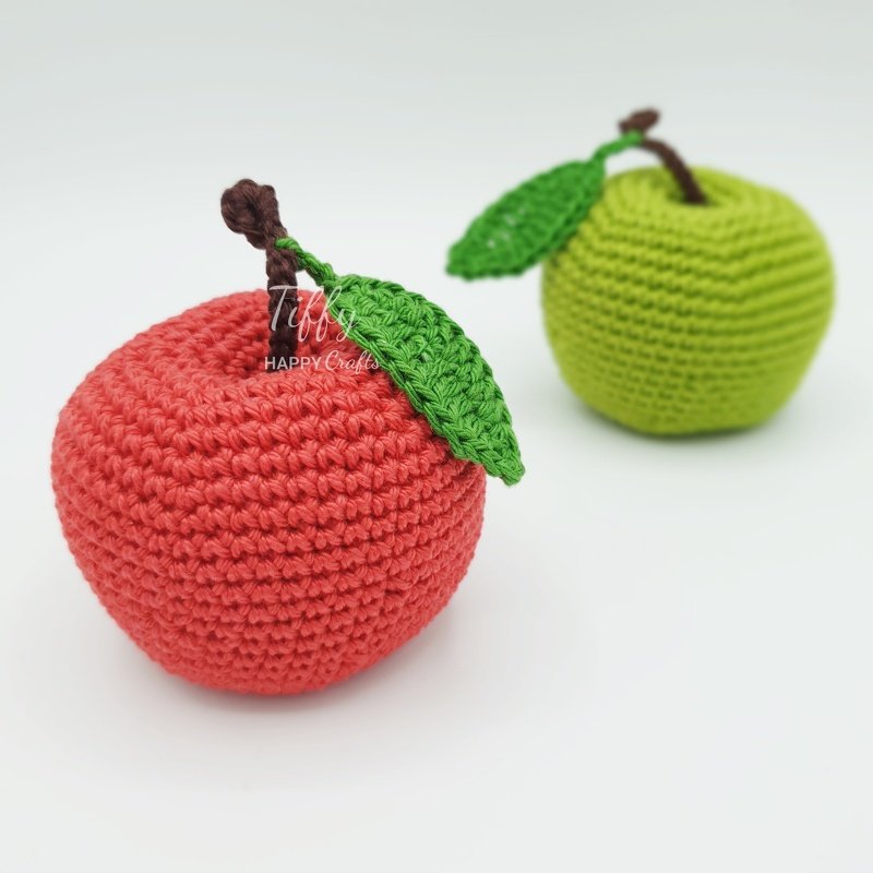 Close-up of a red crocheted apple with stem and leaf, green crocheted apple blurred behind.