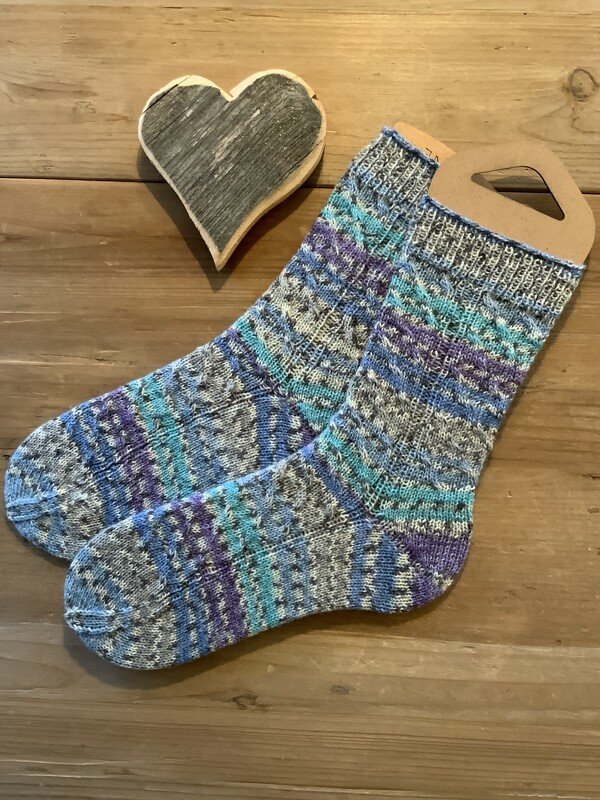 Gestrickte Socken in Blau-, Türkis-, Lila- und Grautönen auf einem Holztisch neben einem Holzherz.