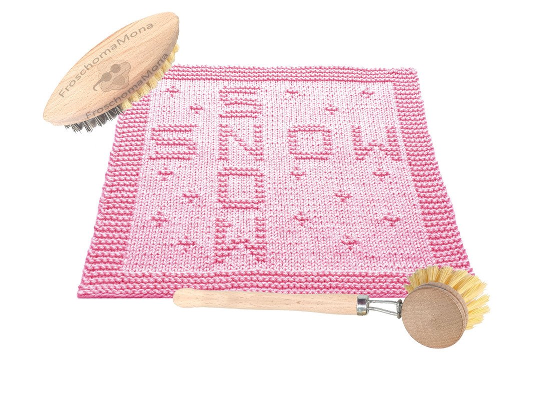 Zartrosa gestrickter Topflappen mit 'SNOW'-Muster und dekorativer Randbordüre, neben zwei Holzbürsten auf weißem Hintergrund