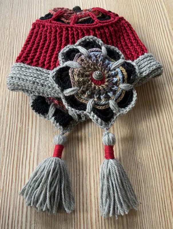 Rote gehäkelte Mütze mit mehrfarbiger Blumenapplikation, grauem Umschlag und zwei grauen Quasten auf hellem Holzuntergrund.