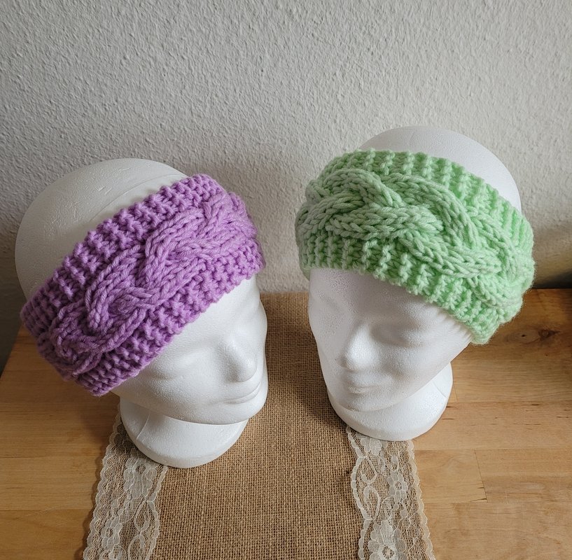 Zwei gestrickte Stirnbänder mit Zopfmuster in Lila und Hellgrün auf weißen Schaufensterköpfen.