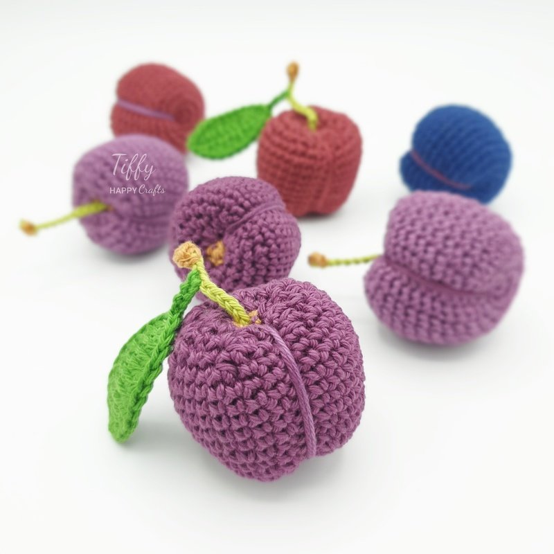 Handgehäkelte pflaumenförmige Amigurumi in Lila, Rot und Blau mit grünen Blättern auf weißem Hintergrund