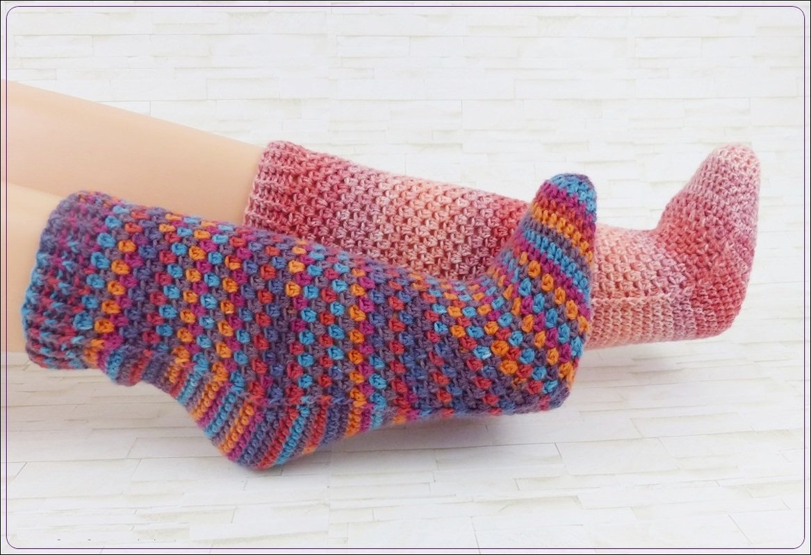 Zwei handgehäkelte, knöchelhohe Socken: eine bunte mit Noppenmuster, die andere rosa im Farbverlauf.