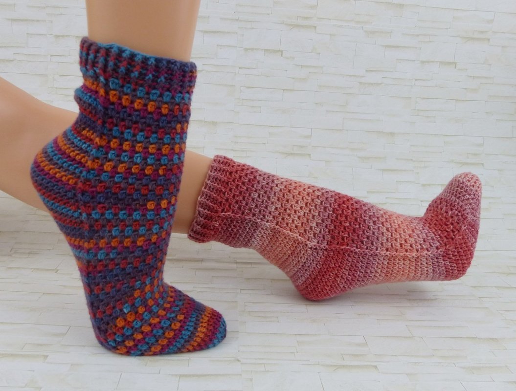 Zwei Füße mit handgehäkelten Socken, eine mehrfarbig gepunktet, die andere in Rot‑ und Rosatönen.
