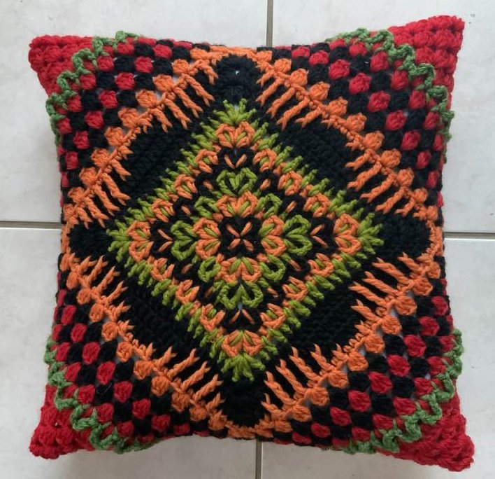 Quadratisches gehäkeltes Kissen in Schwarz, Orange, Grün und Rot mit geometrischem Rautenmuster auf hellem Fliesenboden.