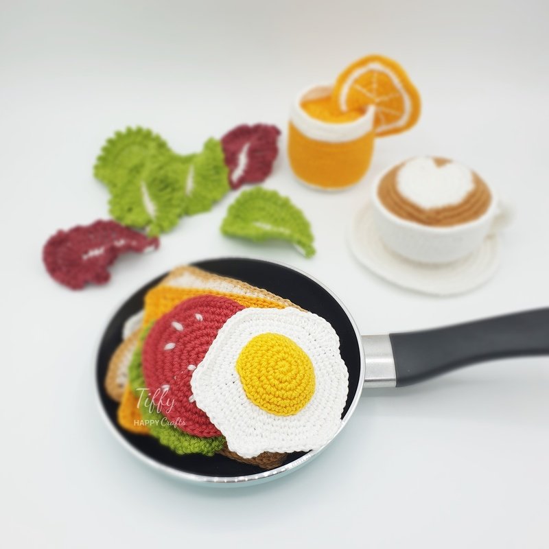 Gehäkeltes Spielzeug-Frühstück: Spiegelei, Sandwich, Salat, Speck, Orangensaft und Kaffee in einer Pfanne auf weißem Hintergrund