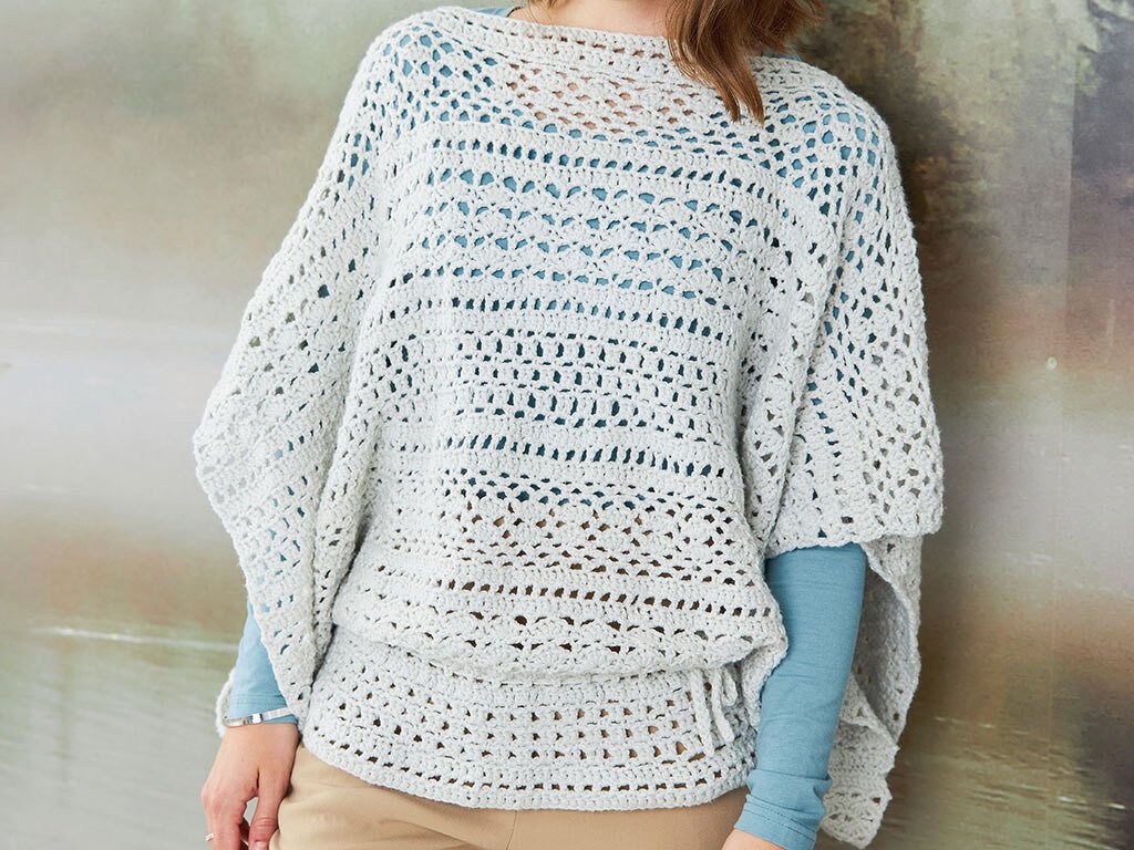 Hellgrauer gehäkelter Poncho mit offener Lochstruktur über blauem Langarmshirt.